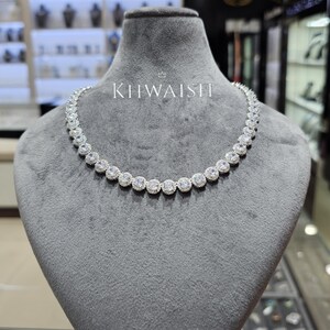 Puede incluir: Un collar de tono plateado que presenta una hilera continua de piedras redondas, transparentes y facetadas, cada una rodeada por un halo de piedras más pequeñas y brillantes. La palabra KHWAISH está impresa en letras blancas sobre el collar.