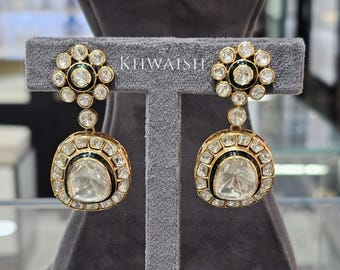 May include: A pair of gold-toned earrings with a floral design at the top and a large, rounded bottom. The earrings are encrusted with clear gemstones and feature a black enamel border. The text "KHWAISH" is visible.