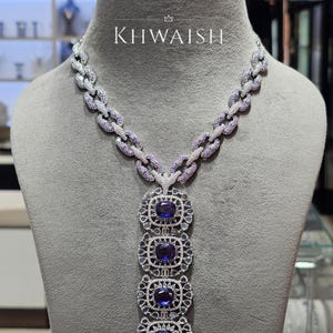 Puede incluir: Un collar con una cadena de eslabones entrelazados incrustados con pequeñas piedras moradas y blancas. Un colgante vertical cuelga del centro, compuesto por cuatro marcos cuadrados, cada uno con una gran piedra azul oscuro y el texto KHWAISH.