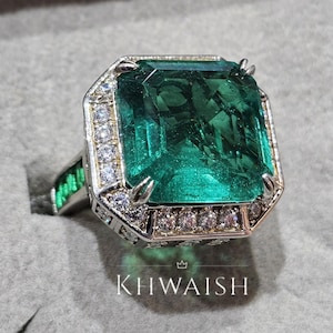 Peut inclure: Une bague en argent ornée d'une grande pierre verte émeraude taillée en émeraude. La bague est entourée de plus petites pierres précieuses claires et possède une bande avec des accents verts. Le texte "KHWAISH" est visible.
