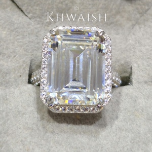 Peut inclure: Une bague en argent ornée d'un grand diamant de taille émeraude, entouré d'un halo de petites pierres. La bague est présentée sur une surface grise. Le mot "KHWAISH" est imprimé en blanc en haut de l'image.