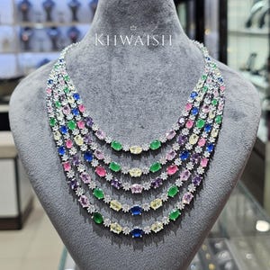 Puede incluir: Un collar de múltiples hilos con cinco filas de piedras preciosas ovaladas multicolores, incluyendo tonos azules, verdes, amarillos, rosas y morados, intercaladas con pequeños cristales transparentes en metal plateado. El texto KHWAISH es visible.