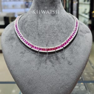 Pode incluir: Um colar com uma fileira de gemas cor-de-rosa de corte quadrado, ladeada por duas filas de pequenas pedras claras numa armação metálica. A palavra KHWAISH está impressa a branco sobre o colar, com um pequeno ícone de coroa acima.