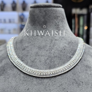 Puede incluir: Un collar de tono plateado que presenta múltiples filas de piedras transparentes de corte baguette dispuestas en una banda densa y continua. La pieza muestra el texto KHWAISH con un pequeño icono de corona encima, centrado sobre el collar.