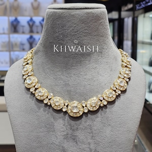 May include: A gold-toned necklace with large, clear gemstones set in a circular pattern. The necklace is displayed on a gray mannequin. The text "KHWAISH" is visible on the mannequin.