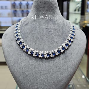 Puede incluir: Un collar de tono plateado con un patrón floral repetitivo de piedras azul oscuro de corte ovalado rodeadas por pequeñas piedras claras. La pieza se exhibe sobre un busto de terciopelo gris e incluye el texto KHWAISH impreso en blanco arriba.