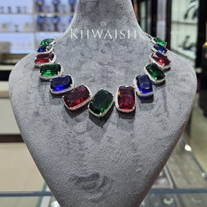 Puede incluir: Un collar con grandes piedras preciosas de corte rectangular en tonos rojo intenso, verde esmeralda y azul real. Cada piedra está enmarcada por un borde de pequeñas piedras transparentes engastadas en pavé. El texto KIIWAISH está impreso en blanco.