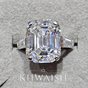 Peut inclure: Une bague en diamant clair de taille émeraude avec des pierres latérales en baguette. La bague est sertie dans un métal argenté et est présentée sur une surface grise. Le nom de la marque "KIIWAISH" est imprimé en bas de l'image.