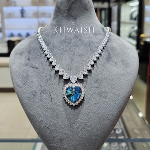 Könnte beinhalten: Eine silberne Halskette mit einem großen, herzförmigen, blauen Edelsteinanhänger. Der Anhänger ist von kleineren, klaren Edelsteinen umgeben. Die Halskette wird auf einer grauen Schaufensterpuppe präsentiert. Der Text "KHWAISH" ist sichtbar.
