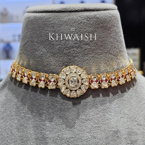 Puede incluir: Un collar gargantilla dorado con un colgante central circular y un patrón repetido de piedras transparentes y rojas. El collar se exhibe sobre una cabeza de maniquí gris. La palabra "KHWAISH" está impresa en blanco.