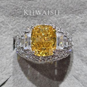 Pode incluir: Um anel luxuoso com um grande diamante amarelo radiante como peça central, ladeado por diamantes brancos retangulares. A banda é cravejada com diamantes brancos menores e brilhantes. A marca "KHWAISH" é exibida acima.
