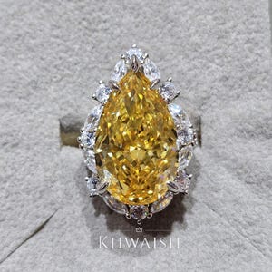 Puede incluir: Anillo con una gran gema amarilla en forma de pera, rodeada de piedras blancas más pequeñas. El anillo está engarzado en un metal plateado o blanco. El nombre de la marca "KIIWAISH" está impreso en la parte inferior.