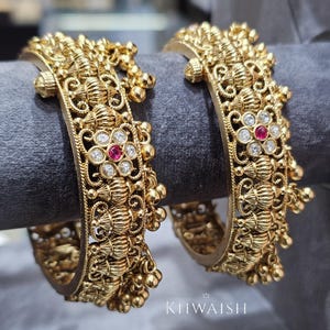 Può includere: Due braccialetti dorati ornati con intricati disegni floreali e in filigrana. Ogni braccialetto presenta un motivo floreale centrale con una gemma rossa circondata da pietre trasparenti. Piccole perle d'oro pendono dai lati. I braccialetti sono esposti su una superficie grigia.
