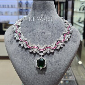 Puede incluir: Un collar de tono plateado con una gran piedra central de color verde oscuro en talla cojín, rodeada de piedras claras redondas y baguette. La cadena muestra un patrón ondulado con pequeñas piedras de color rosa vibrante y cristales baguette. El texto KHWAISH es visible sobre el collar.