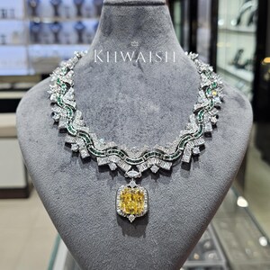 Puede incluir: Un collar con una gran piedra central amarilla de corte cojín rodeada de diamantes claros en corte baguette y redondos, acentuado con pequeñas piedras verdes en un patrón ondulado. El texto KIIWAISHI es visible sobre el collar en el expositor.