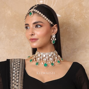 Może przedstawiać: Zdobiony, złoty zestaw biżuterii z naszyjnikiem typu choker, kolczykami i nakryciem głowy, wszystkie zdobione przezroczystymi i szmaragdowo-zielonymi kamieniami szlachetnymi. Naszyjnik i nakrycie głowy mają geometryczny wzór. Widoczne jest słowo "KHWAISH".