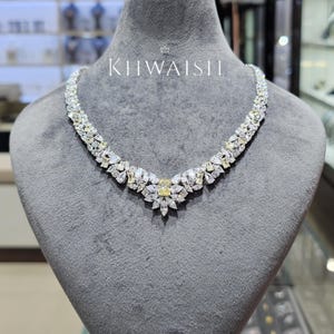 Puede incluir: Un collar de tono plateado con una piedra rectangular amarilla central rodeada de piedras transparentes en forma de pera y corte marquesa. El diseño continúa con piedras transparentes y amarillas alternas a lo largo de la cadena. El texto KHWAISH es visible.
