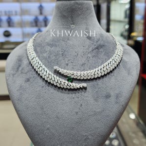 Könnte beinhalten: Eine Diamantkette mit blattartigem Design und einem grünen Edelstein. Die Kette wird auf einer grauen Schaufensterpuppe präsentiert. Der Markenname "KHWAISH" ist in weißer Schrift zu sehen.