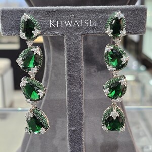 May include: Elegant drop earrings featuring large, pear-shaped emerald green gemstones. The earrings are accented with smaller green stones and sparkling white crystals. The brand name "KHWAISH" is displayed above the earrings.