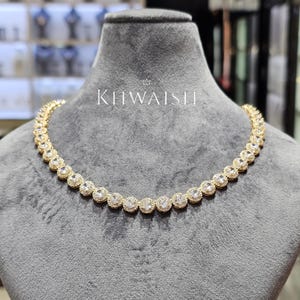 Puede incluir: Un collar de tono dorado con una fila continua de eslabones circulares, cada uno con una piedra central transparente rodeada por un halo de piedras más pequeñas. La palabra KHWAISH está impresa en texto blanco sobre el centro del collar.