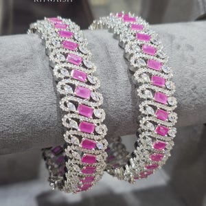 Peut inclure: Deux bracelets rigides argentés ornés d'une rangée centrale de pierres roses rectangulaires flanquées de rangées de pierres claires taille marquise. La monture métallique est incrustée de petites pierres claires. Le texte KHWAISH est visible.
