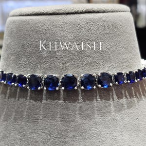 Puede incluir: Un collar tipo tenis de tono plateado que presenta una hilera continua de piedras redondas facetadas de color azul profundo. La palabra KHWAISH está impresa en blanco sobre el collar. La joya se exhibe en un busto cilíndrico gris texturizado.
