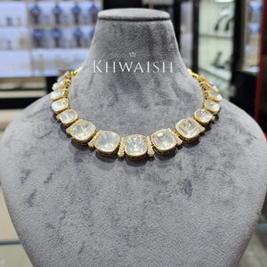 May include: A gold necklace featuring a series of square-cut, clear, faceted stones set in a row. The metal settings are accented with small, sparkling stones. The word KHWAISH is printed in white above the necklace on the gray display bust.
