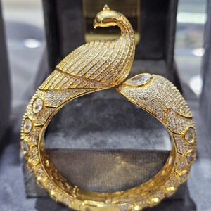 Könnte beinhalten: Ein goldfarbenes Armband mit einem Pfauenmotiv, das mit zahlreichen kleinen, klaren Edelsteinen besetzt ist. Der Kopf und der Körper des Pfaus sind mit den Edelsteinen detailliert. Das Armband trägt oben das Wort "Khwaish".