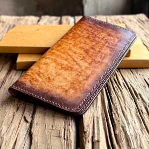 Puede incluir: Cartera de cuero marrón con acabado envejecido. La cartera presenta un borde cosido y un aspecto texturizado. El cuero tiene un degradado de colores, desde el marrón claro hasta el marrón oscuro. Hecho a mano.