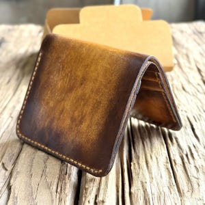 Puede incluir: Una cartera de cuero marrón con un acabado envejecido. La cartera está abierta, revelando el interior. Los bordes están cosidos con un hilo en contraste. Una caja de cartón está en el fondo. La cartera está sobre una superficie de madera.