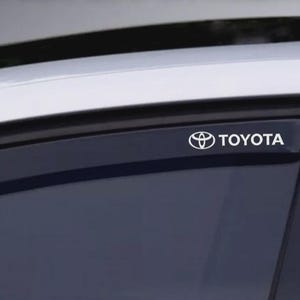 May include: A close-up of a car window with a white Toyota logo and text. The logo is a stylized emblem with the brand name in a sans-serif font. The window is tinted, and the car's roof is visible.