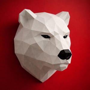 Op de afbeelding: Een witte, geometrische ijsberenhoofdsculptuur met een zwarte neus en ogen. De sculptuur is gemonteerd op een rode achtergrond, wat een opvallend contrast creëert. De vorm van de beer is opgebouwd uit meerdere driehoekige vlakken.