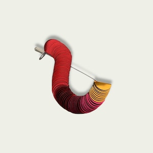 Könnte beinhalten: Eine einzigartige Brosche mit geschwungenem Design aus geschichteten, kreisförmigen Elementen. Die Brosche wechselt von Rot zu Gelb, mit einem silbernen Nadelverschluss.