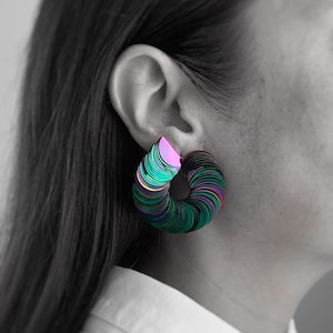 May include: A close-up of a person wearing a large, iridescent sequin earring. The earring is a spiral shape with layers of green, purple, and pink sequins. The background is black and white, with the person's ear and part of their hair and shirt visible.