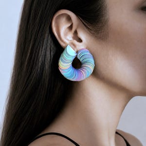 May include: A circular hoop earring featuring a stacked, layered design with a gradient of iridescent pastel colors including light blue, pink, and yellow. The earring has a smooth, curved structure that forms a continuous loop shape on the earlobe.