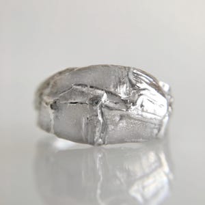 May include: A silver ring with a textured, irregular surface. The ring has a unique, organic shape, reflecting light and creating highlights and shadows. The design appears to be handmade, with visible imperfections adding to its character. The ring is displayed on a reflective surface.