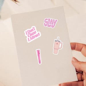 May include: A set of three stickers featuring a pink text sticker that reads I Don't Chase I Attract, a pink wavy text sticker that reads SLAY, and a pink tumbler with a straw and a pink lip treatment tube with the text rhode printed on it.