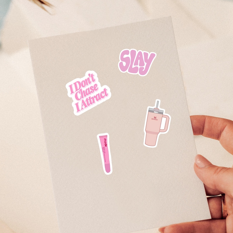 May include: A set of three stickers featuring a pink text sticker that reads I Don't Chase I Attract, a pink wavy text sticker that reads SLAY, and a pink tumbler with a straw and a pink lip treatment tube with the text rhode printed on it.
