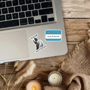 May include: Laptop with stickers. One sticker says "HELLO, I'M easily distracted" in a blue and white name tag design. Another sticker features a black and white image of a man with the words "I KNOW NOTHING".