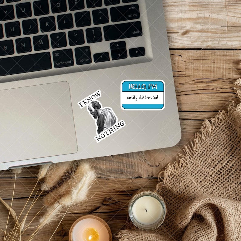 May include: Laptop with stickers. One sticker says "HELLO, I'M easily distracted" in a blue and white name tag design. Another sticker features a black and white image of a man with the words "I KNOW NOTHING".