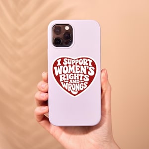 May include: A light purple phone case with a red heart-shaped sticker. The sticker reads "I SUPPORT WOMEN'S RIGHTS AND WRONGS" in a retro font. The phone case is held in a person's hand.