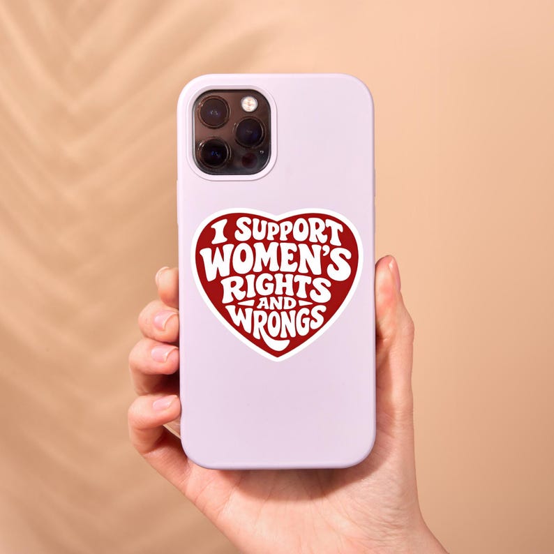 May include: A light purple phone case with a red heart-shaped sticker. The sticker reads "I SUPPORT WOMEN'S RIGHTS AND WRONGS" in a retro font. The phone case is held in a person's hand.