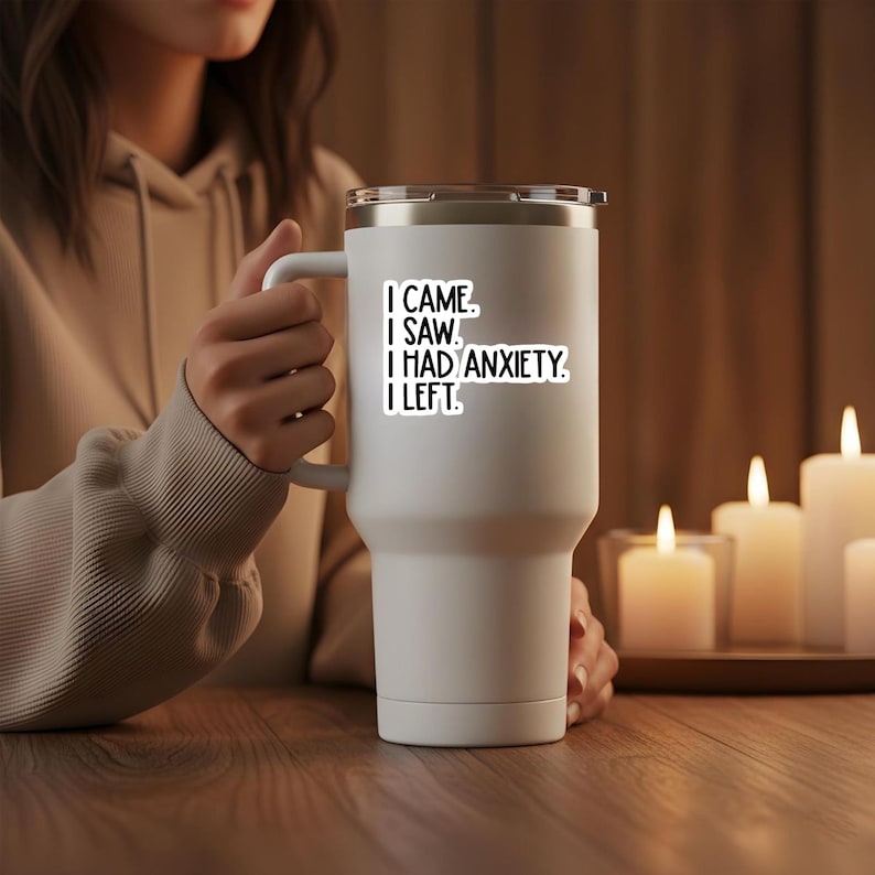 May include: A white insulated tumbler with a handle and a black lid. The tumbler has a sticker with the text "I CAME. I SAW. I HAD ANXIETY. I LEFT." The tumbler is held by a person.