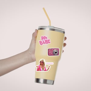 May include: A light yellow stainless steel tumbler with a lid and a yellow straw. The tumbler features three stickers: a pink 90s Babe graphic, a pink digital camera, and a sticker of a person in pink clothing with the text Fabulous.