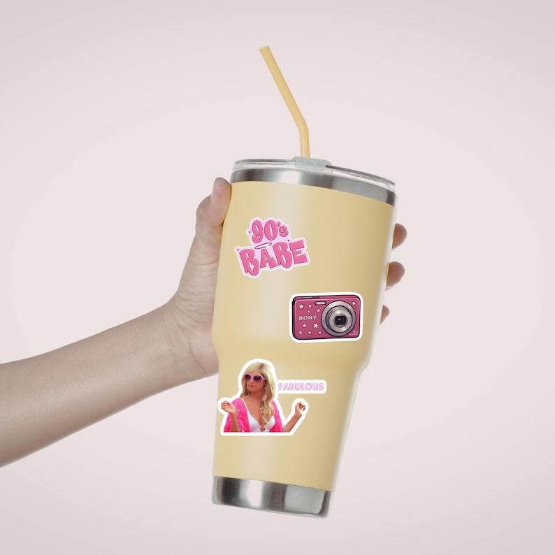 May include: A light yellow stainless steel tumbler with a lid and a yellow straw. The tumbler features three stickers: a pink 90s Babe graphic, a pink digital camera, and a sticker of a person in pink clothing with the text Fabulous.