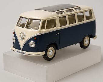 May include: A miniature Volkswagen bus model in cream, navy blue, and gray. The model features the Volkswagen logo on the front and the word "VOLKSWAGEN" below it. The bus has a sunroof and is displayed on a white rectangular base.