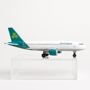 May include: A white and teal model airplane with the Aer Lingus logo on the side and a shamrock on the tail. The toy airplane has two teal engines and is displayed on a clear acrylic stand.