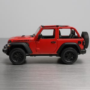 May include: A red Jeep Rubicon model with black wheels and accents. The toy car has a spare tire on the back and the word "RUBICON" is visible on the hood. The vehicle is a two-door model.