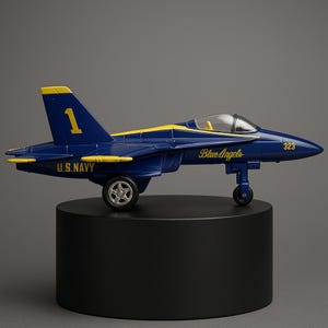 May include: A blue and yellow toy jet plane, labeled "U.S. Navy" and "Blue Angels," sits on a black stand. The plane has the number "1" on the tail and "323" on the side. The jet has a clear cockpit and wheels.