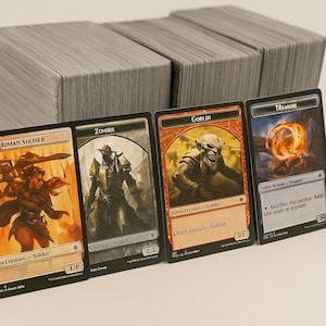 May include: Four Magic: The Gathering trading cards are displayed, featuring fantasy art of a human soldier, a zombie, a goblin, and a treasure. The cards are in front of stacks of gray card stock. The goblin card has the text "Goblin" and "1/1".